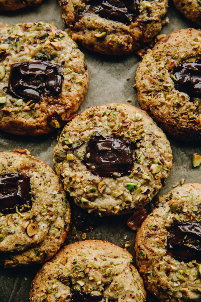 A flat lay shot shows close up of freshly baked pistachio and chocolate cookies revealing a gooey chocolate centre and pistachio crumb around the edge. 