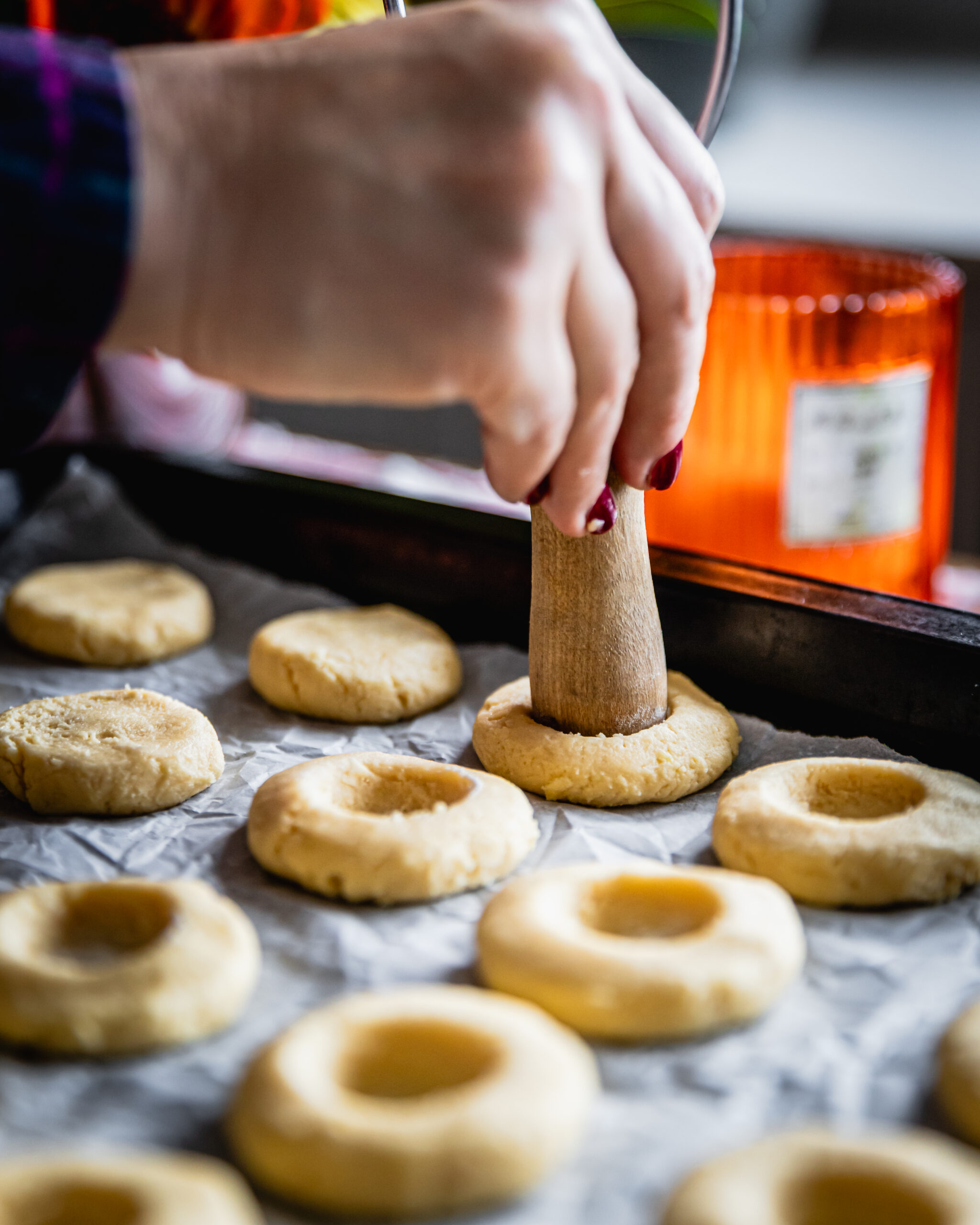 Strawberry jam drop cookies are having holes put in them by a wooden handle. They are on a baking tray with white baking paper. Many of the unbaked cookies already have holes in them.