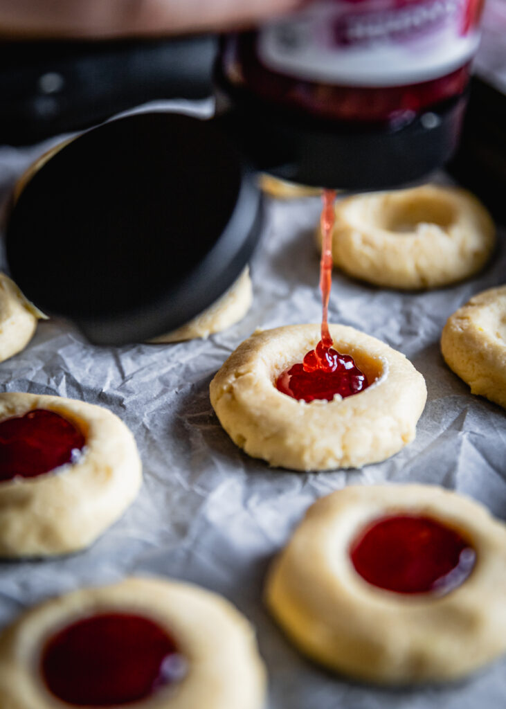 Unbaked strawberry jam drop biscuits are being filled with strawberry jam poured from a bottle.