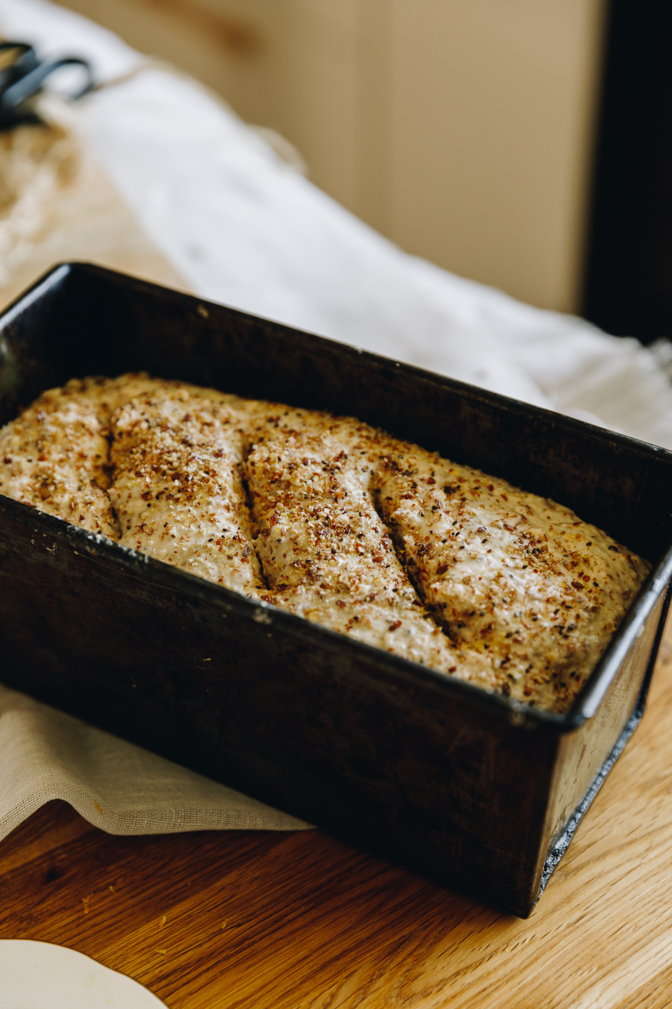 Kneaded Seeded Oat Bread is in a black tin sitting on a wooden table. Sprinkles of fine seeds are on top of the loaf with three slits cut in to it.
