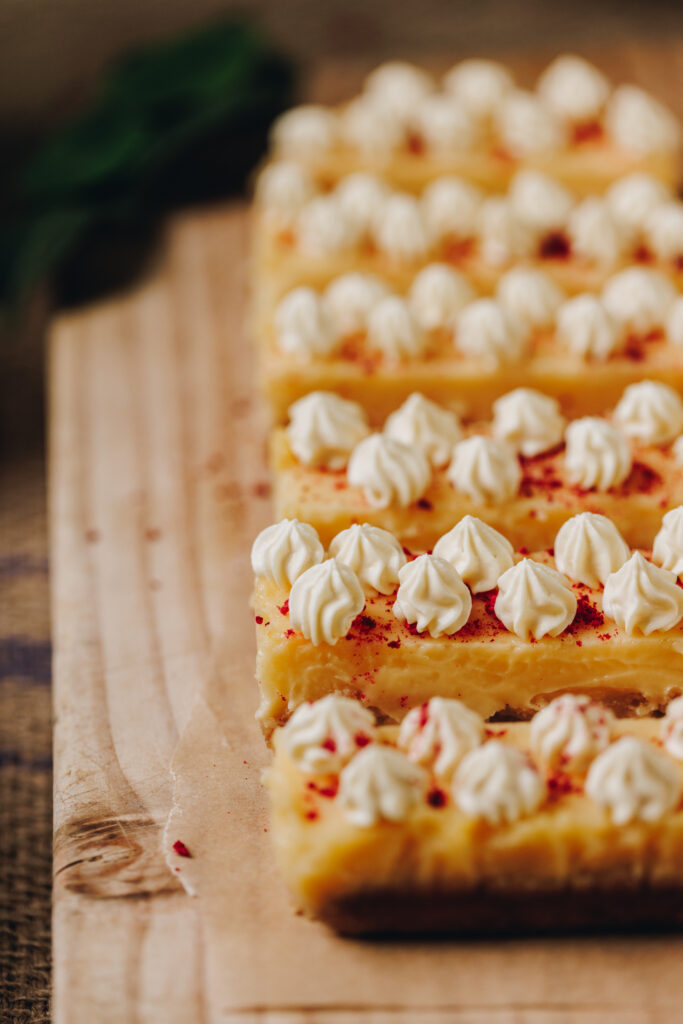 Lemon Bars sit on a wooden board. They are piped with white chocolate yogurt cream and sprinkled with freeze-dried raspberries.