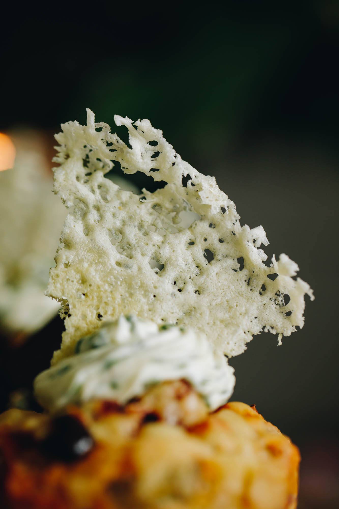 A close shot shows a triangle shaped parmesan wafer crisp on top of a savoury muffin. It is anchored by a herbed cream cheese that is on the centre of the muffin. 