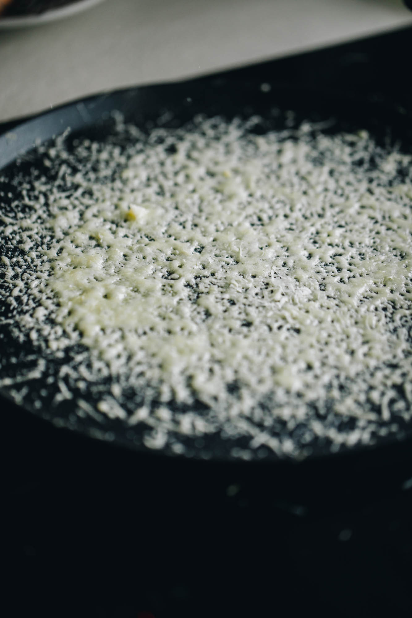 A black frying pan is on a black stove top. IN the pan is grated Parmesan in a round shape. 