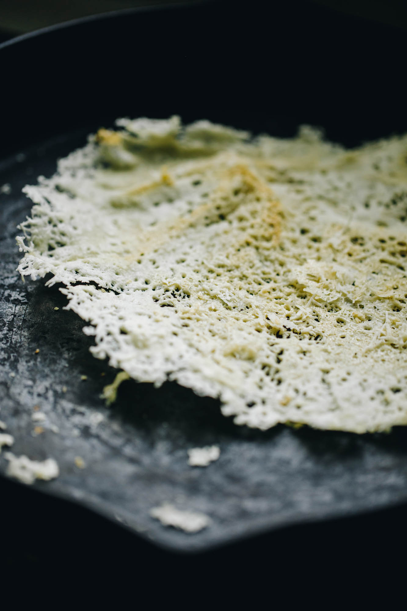 A black frying pan is on a black stove top. In the pan is grated Parmesan in a round shape. The cheese is crisp and is starting to lift off the pan.