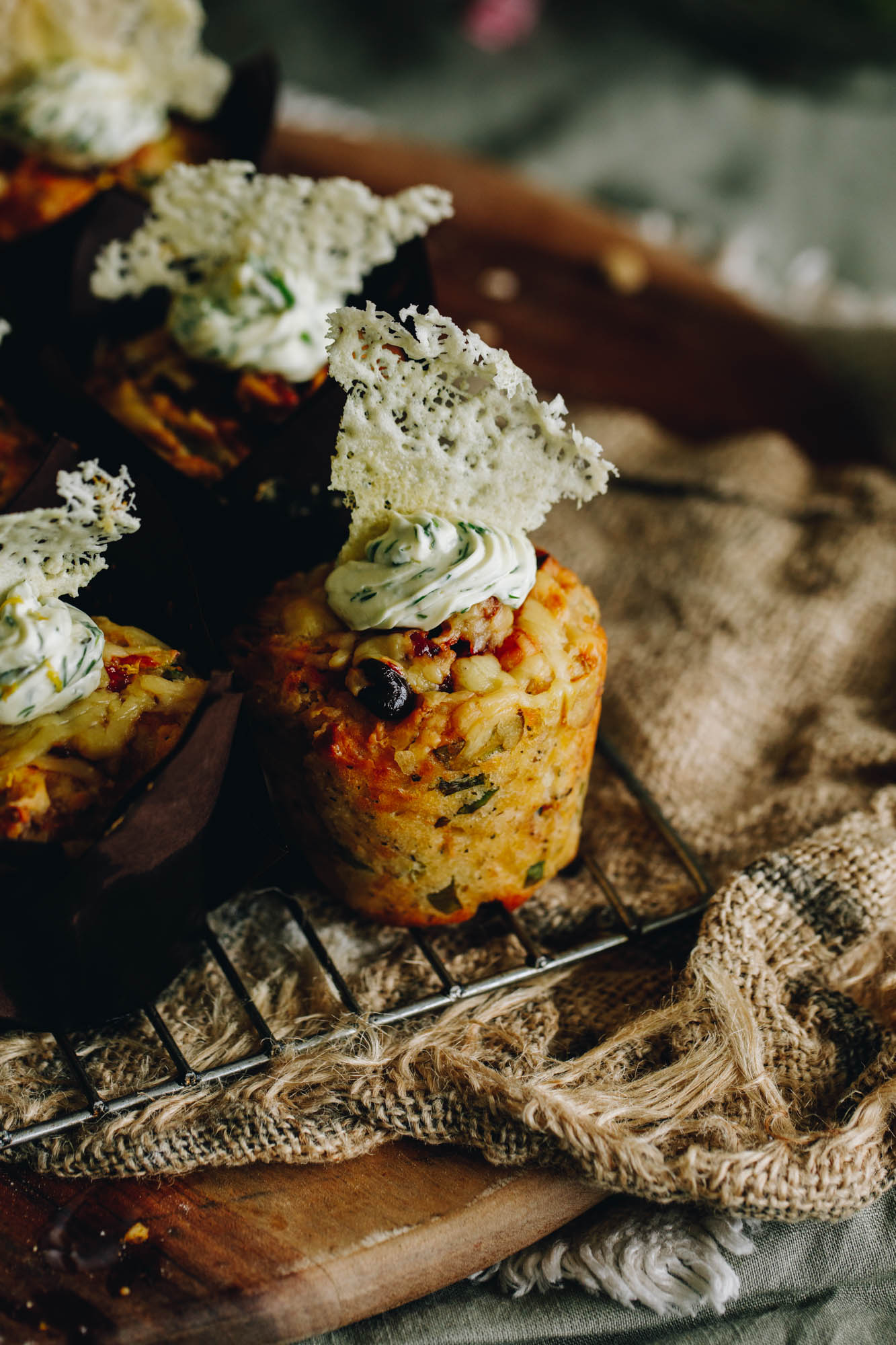 Savoury muffins are on a wire cooling rack on a wooden board with natural fabric. On each of the muffins is a herby cream cheese piped in to the centre and Parmesan Wafer Crisps are on top of each one. 