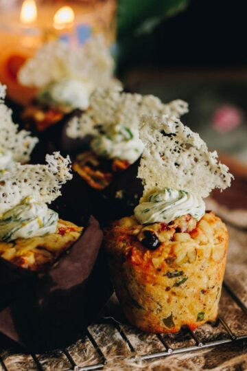 Freshly baked cheesy bacon savoury muffins are sitting on a cooling rack. One of the muffins has no case showing the whole muffin with cream cheese topping and a parmesan crisp on top.