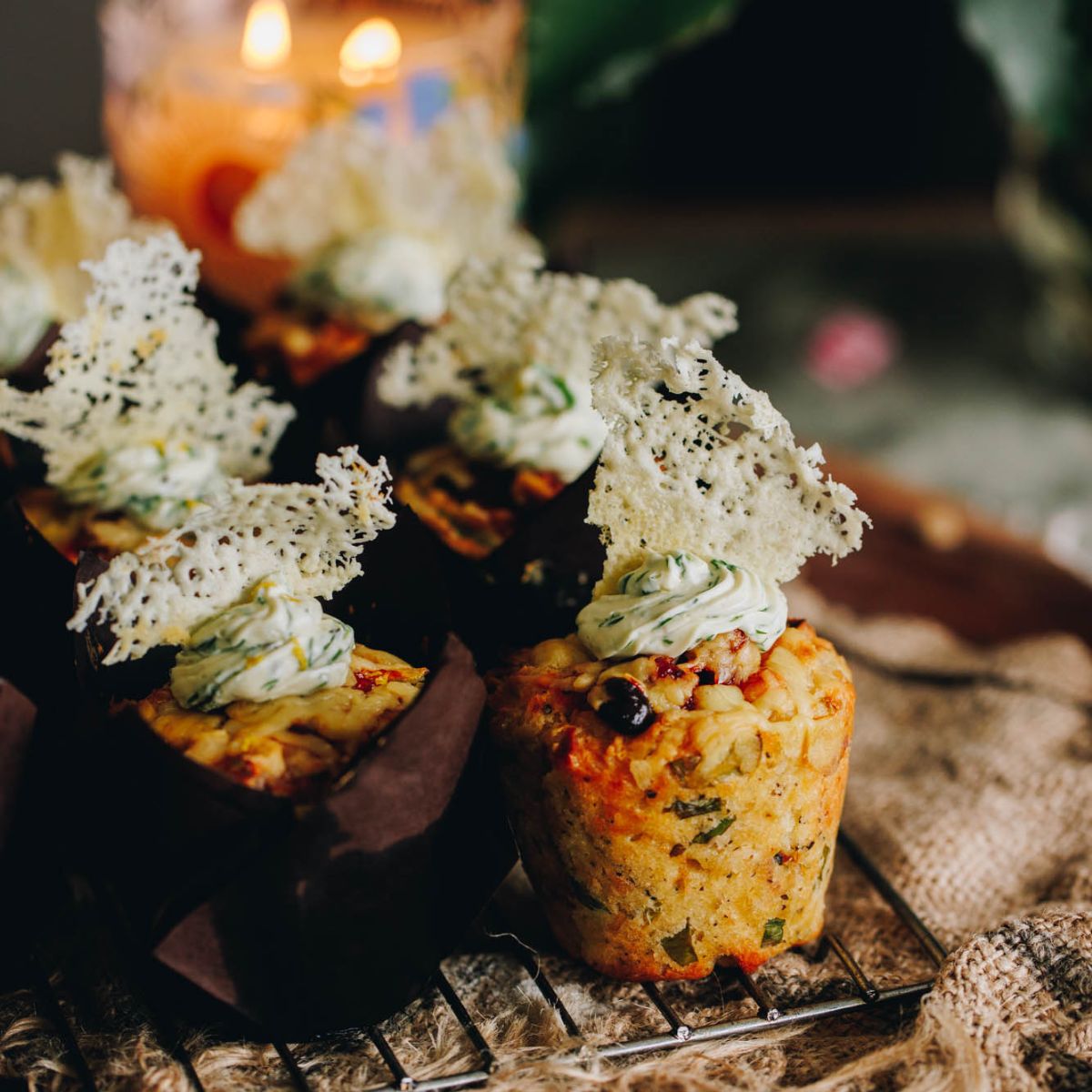 Freshly baked cheesy bacon savoury muffins are sitting on a cooling rack. One of the muffins has no case showing the whole muffin with cream cheese topping and a parmesan crisp on top.