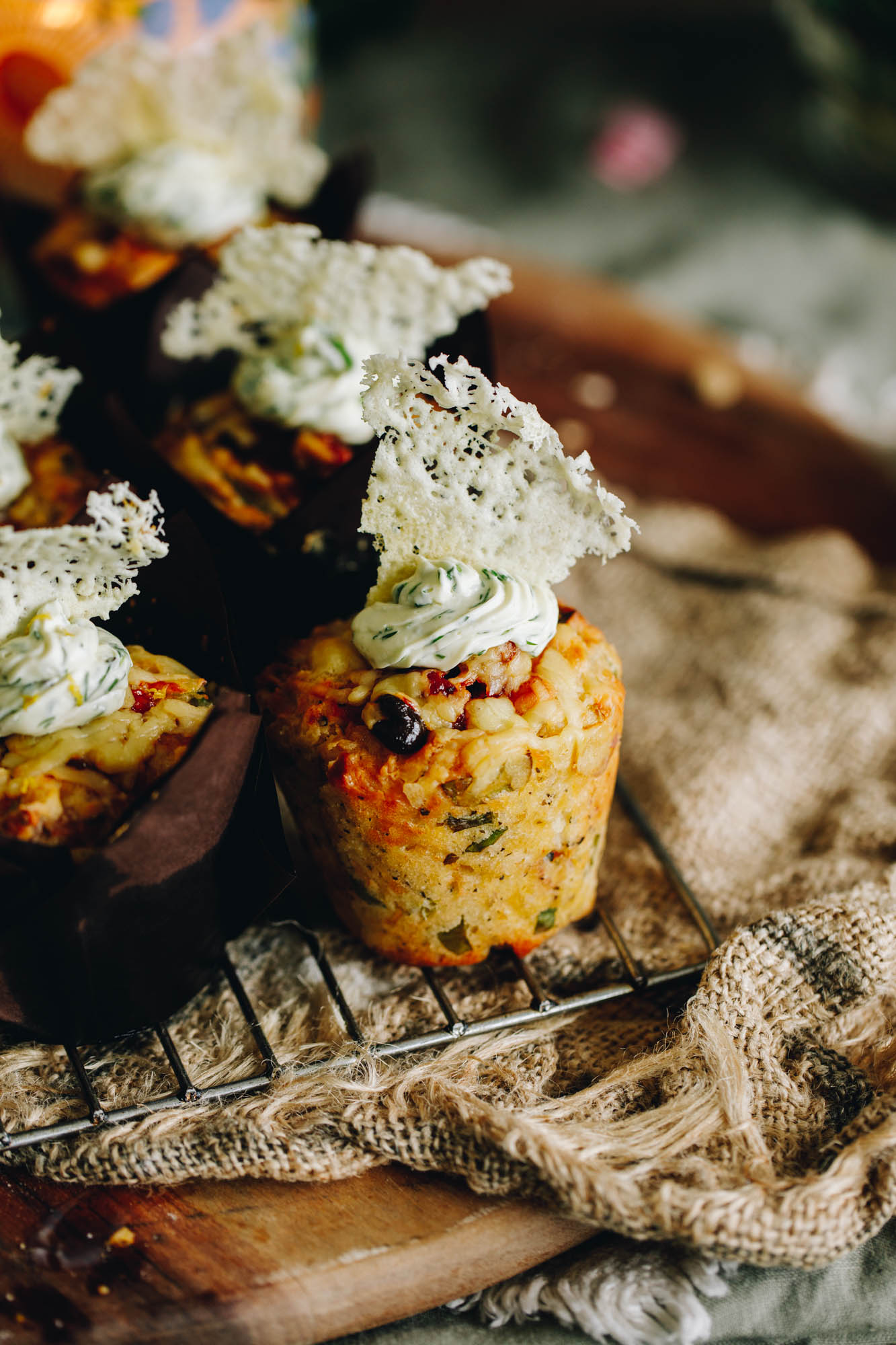 Muffins sit on a cooling rack on top of a wooden board. One muffin is unwrapped and reveals the muffin with cream cheese frosting and paramsen crisp. 