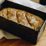 Unbaked No-knead Seeded Oat Loaf is in a black tin sitting on a wooden table. Sprinkles of fine seeds are on top of the loaf with three slits cut in to it.