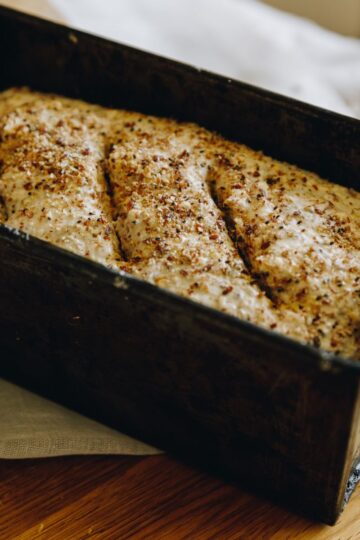 Unbaked No-knead Seeded Oat Loaf is in a black tin sitting on a wooden table. Sprinkles of fine seeds are on top of the loaf with three slits cut in to it.