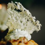 A close up shot of a parmesan wafer is shown, it is sitting on top of herbed cream, on a muffin. The wafer is rustic.