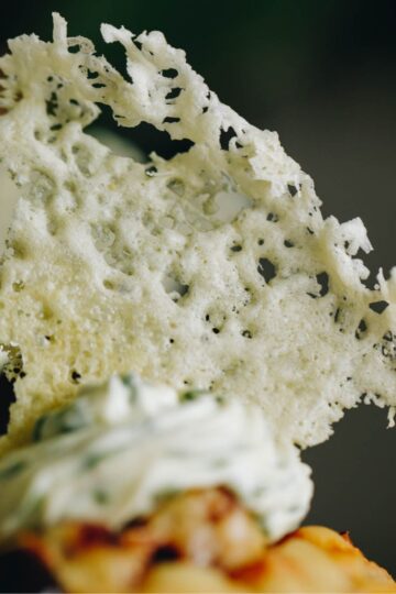 A close up shot of a parmesan wafer is shown, it is sitting on top of herbed cream, on a muffin. The wafer is rustic.