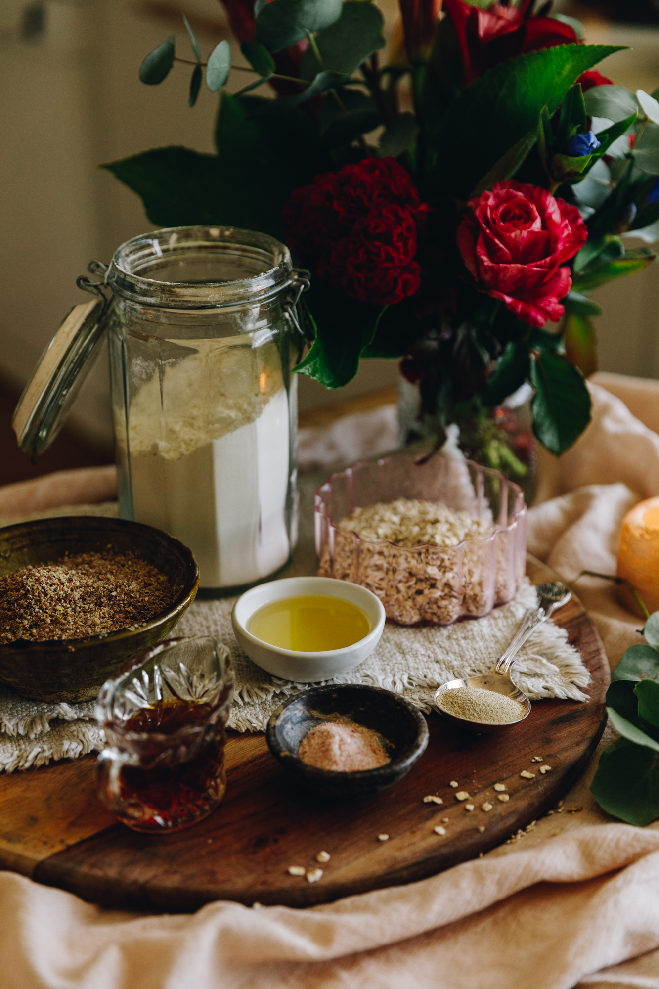 The ingredients for the no-knead-seeded loaf are in different vintage bowls and are sitting on a wooden board. A amber candle and pink flowers are behind the ingredients.