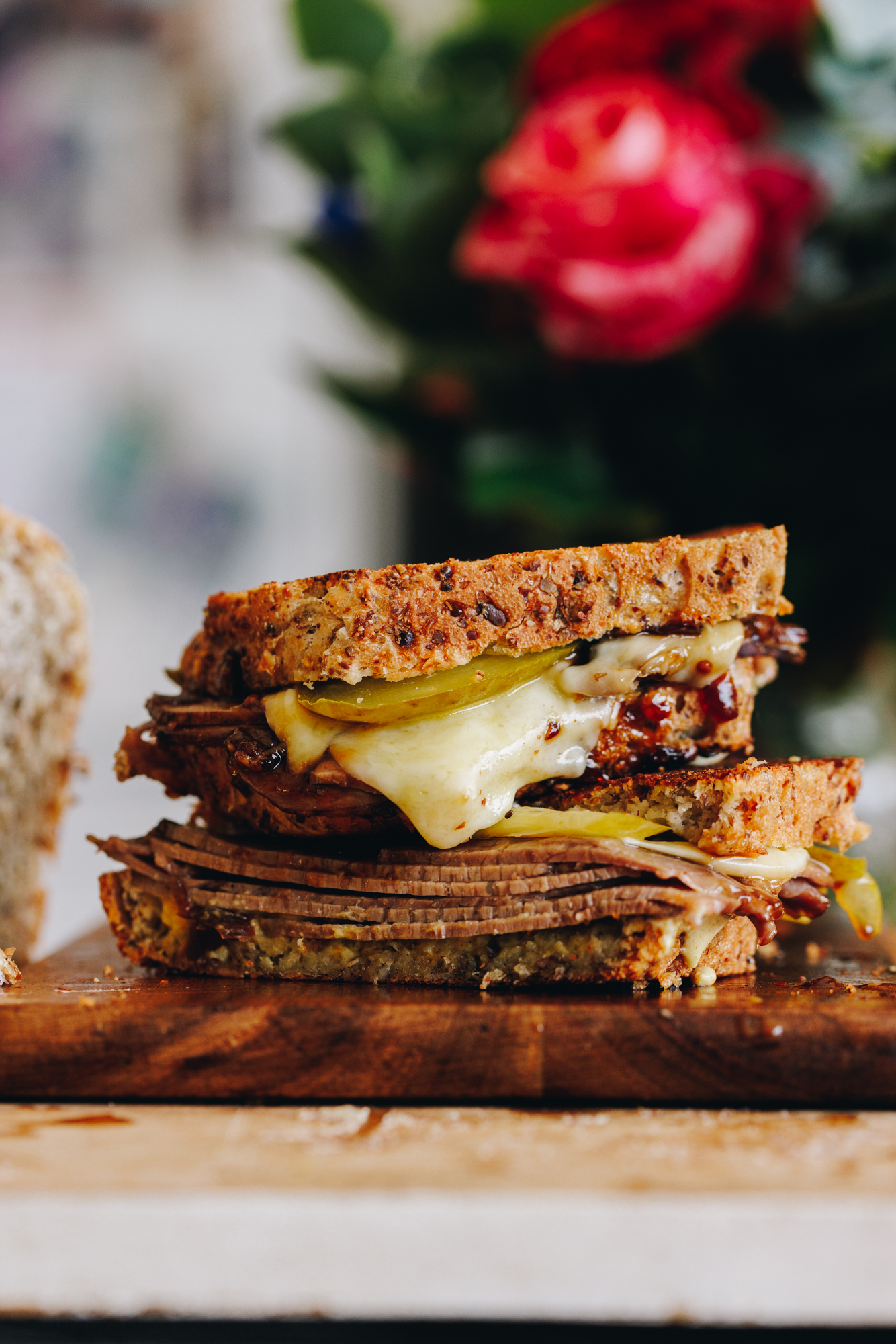 A delicious toasted sandwich is on a wooden board. It has layers of folded meat, pickles and gooey cheese oozing out of it. Pink flowers are in the background.