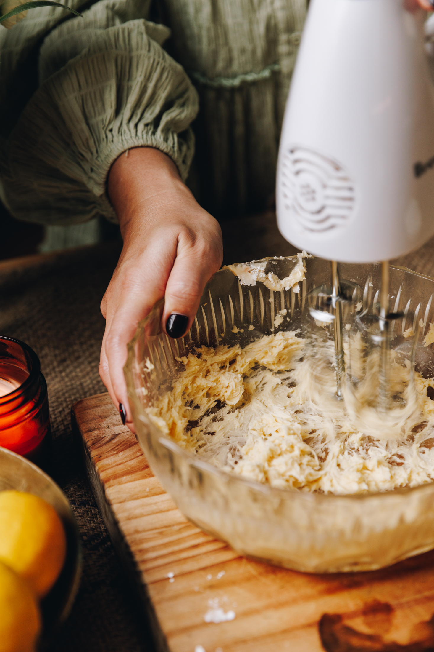 Whipping butter and sugar is in a glass bowl that is sitting on a wooden board. A hand rests on the side of the bowl.