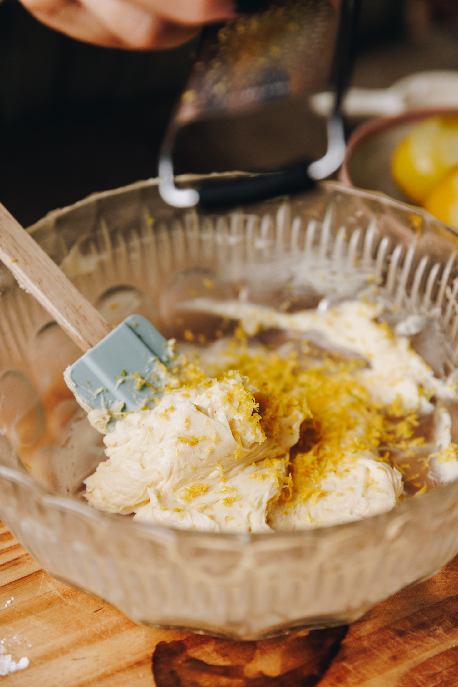 A vintage bowl sits on a wooden table. In the bowl is whipped butter and lemon zest is being grated in to it.