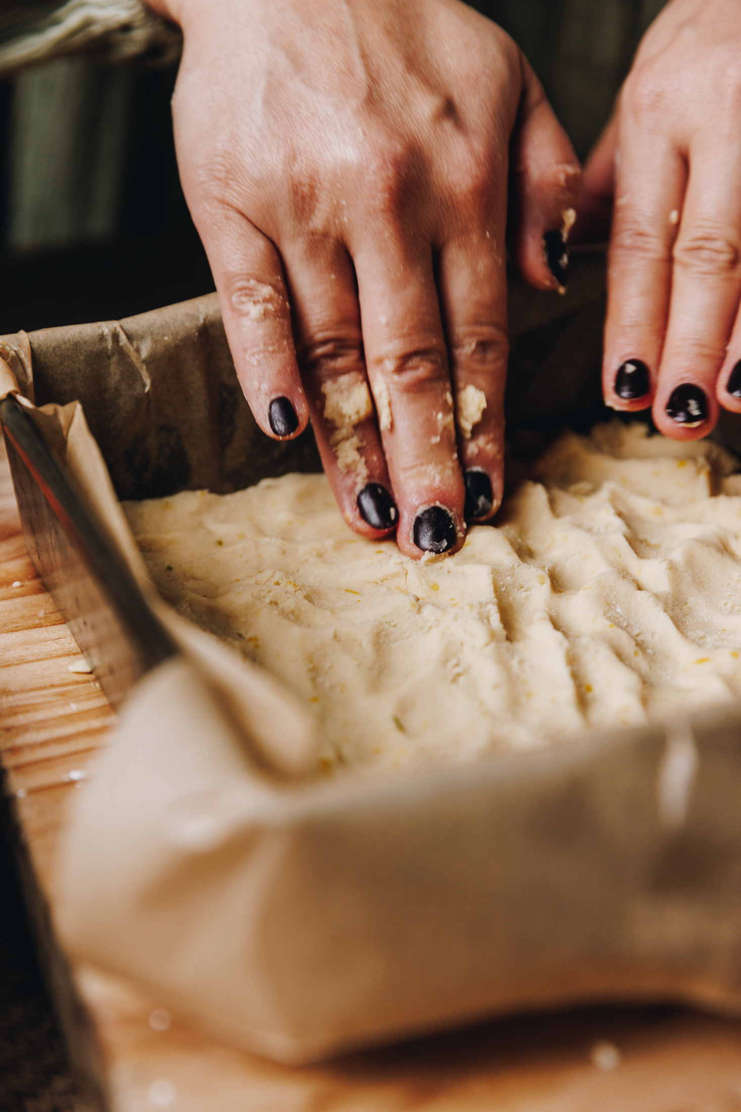 On a wooden table is a baking tin lined with baking paper. The buttery base is being pressed into it with two hands. 