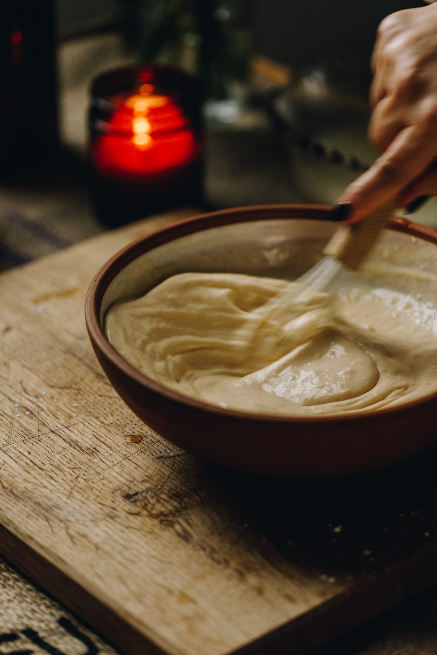 A ceramic bowl is on a wooden board, in the bowl is a lemon custard that is being stirred. An amber candle burns in the background. 