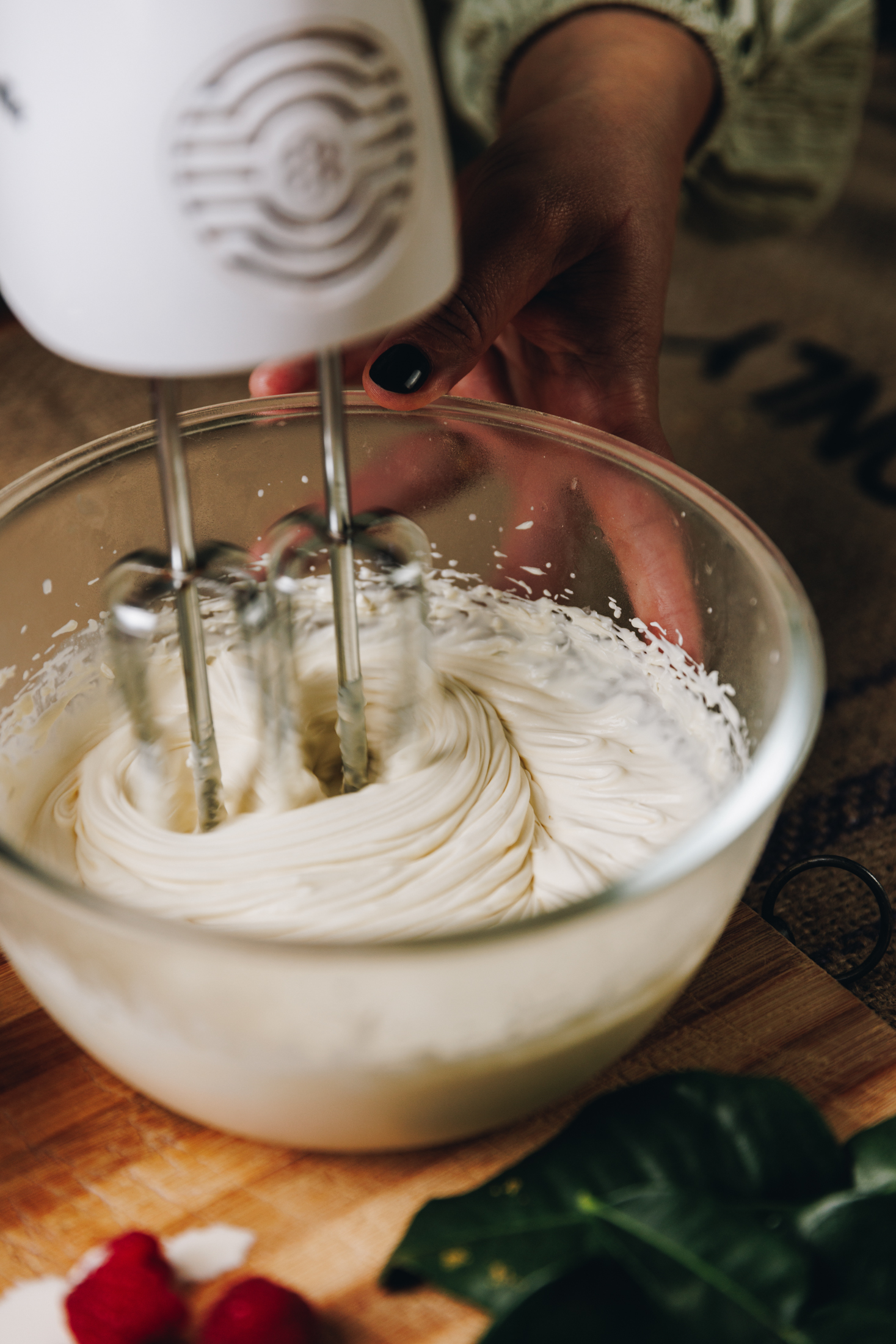 A glass bowl is sitting on a wooden board. In the bowl is cream being whipped with a white hand mixer.