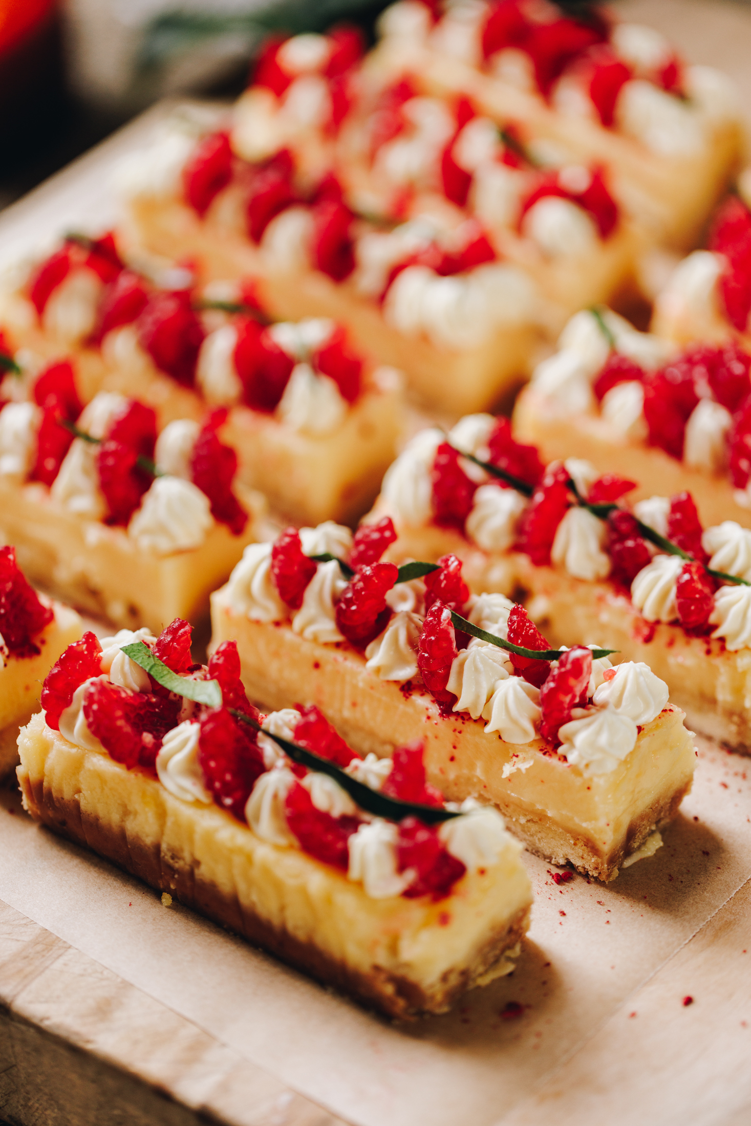 On a wooden board is lemon bars that are decorated with piped whipped cream, slices of fresh raspberries and slivers of lime leaves.