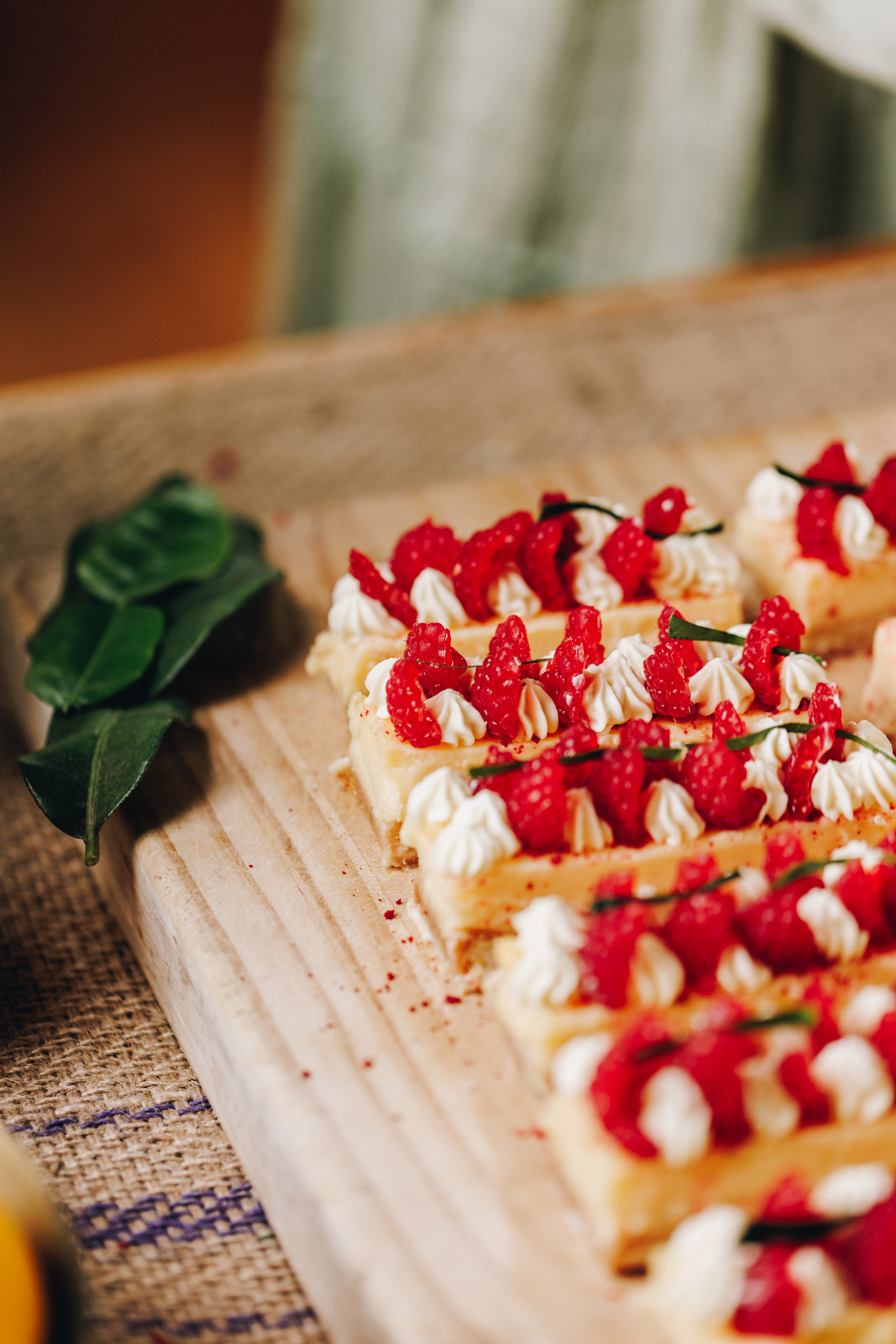 On a wooden board is lemon bars that are decorated with piped whipped cream, slices of fresh raspberries and slivers of lime leaves.