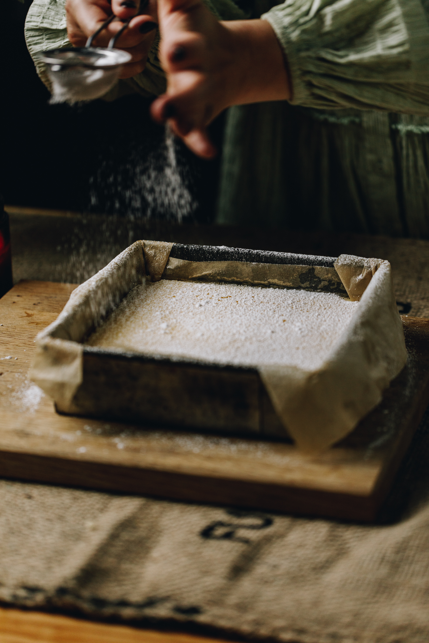 A wooden board is on a wooden table. On the board is a tin with freshly baked lemon bars. The slice is being dusted with icing sugar.