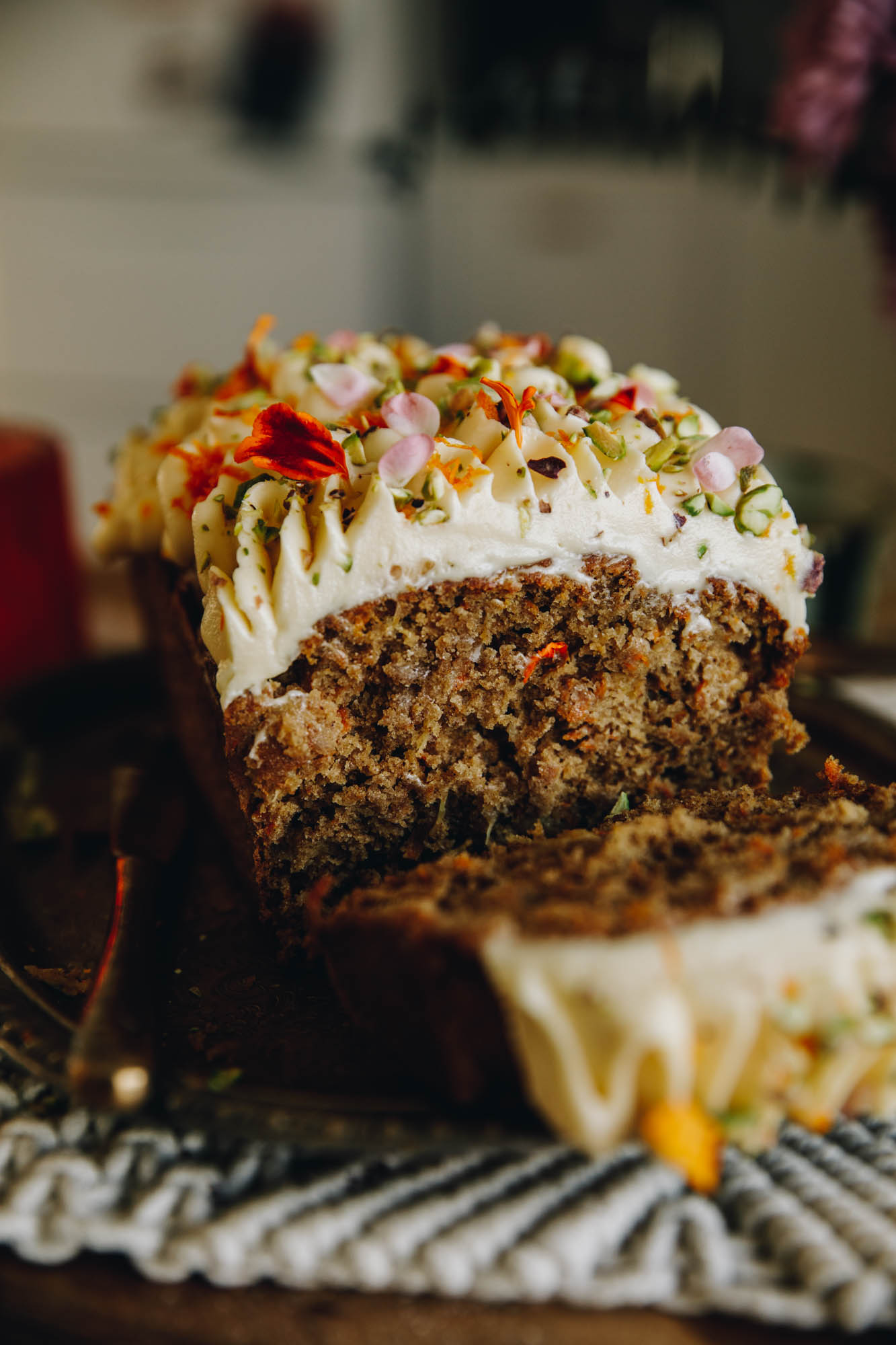 On a wooden table sits a cream weaved mat. On the mat is a freshly bake and decorated Carrot Cake Loaf that has been cut open. It has been decorated with swirls of cream cheese frosting and is topped with chopped pistachios and edible flower petals. 