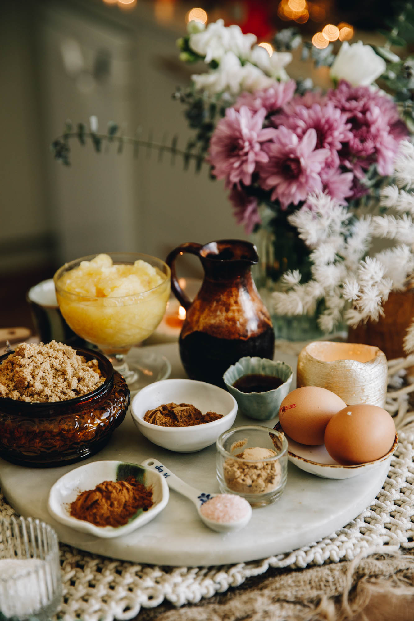 On a white stone sits vintage bowls and small plates that have cake ingredients on it. There is eggs, spices, pineapple and sugar in view. Purple and white flowers are in the background. 