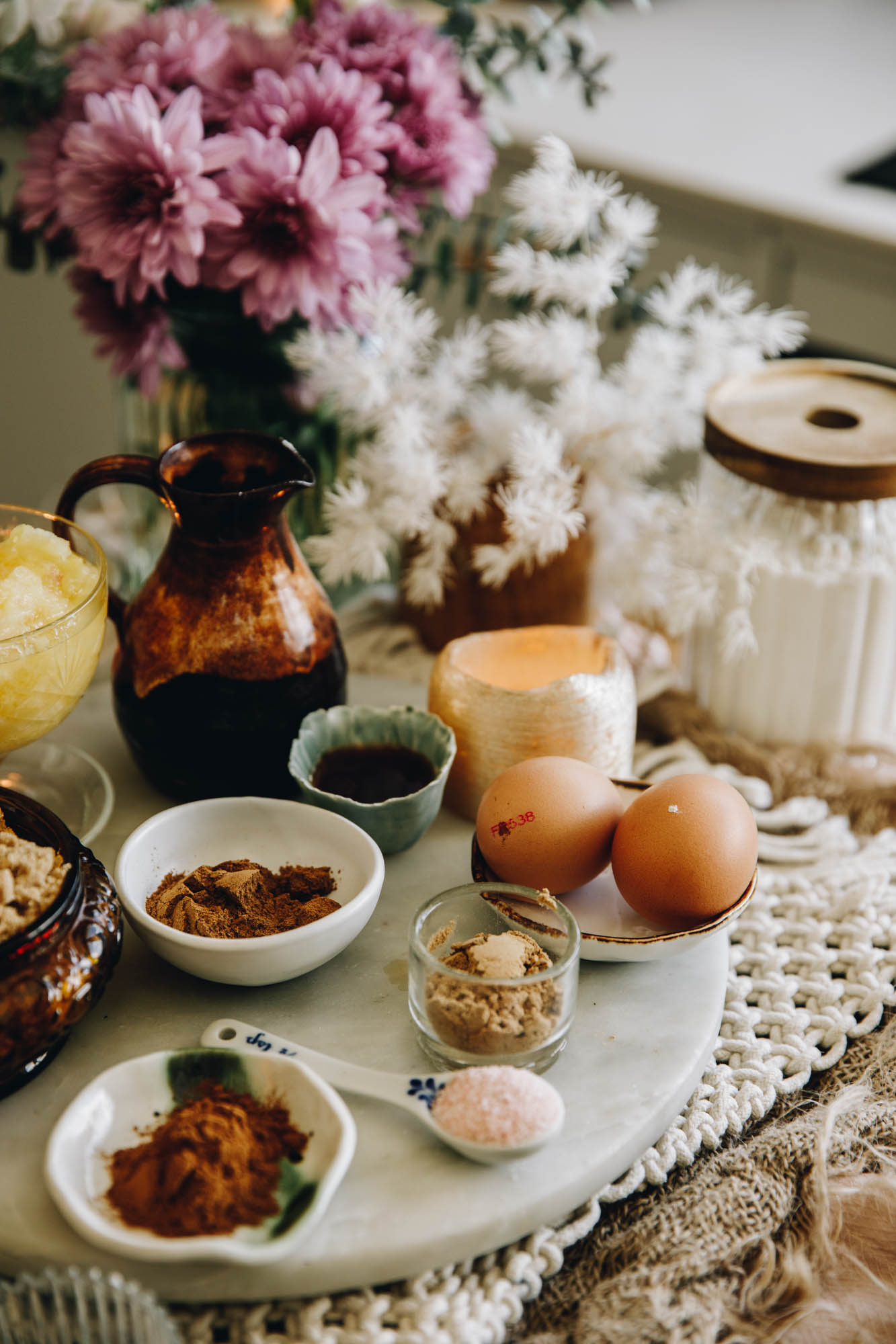 On a white stone sits vintage bowls and small plates that have cake ingredients on it. There is eggs, spices, pineapple and sugar in view. Purple and white flowers are in the background.