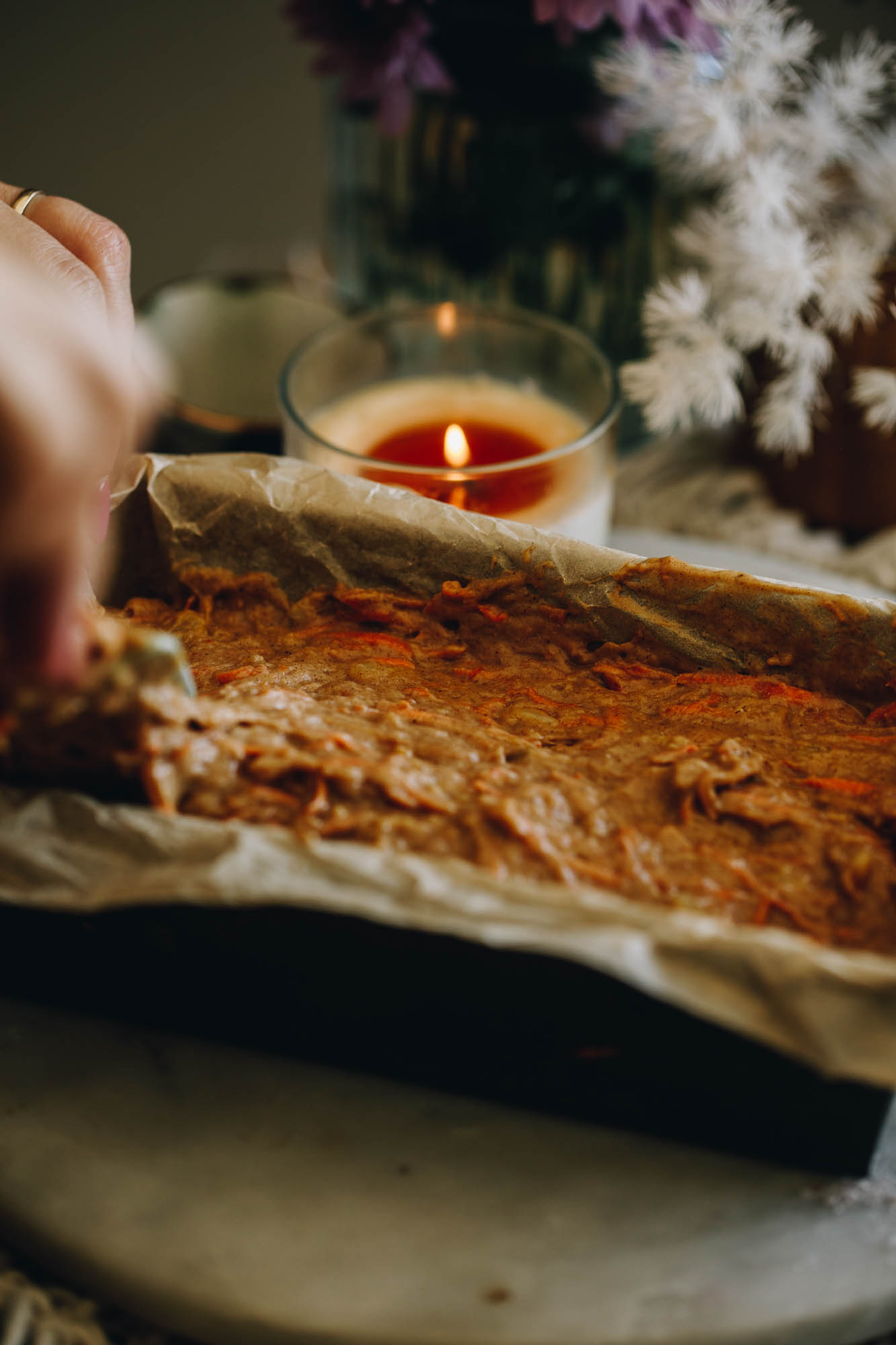 A loaf tin lined with brown baking paper is sitting on a white stone. In the loaf tin is unbaked carrot cake loaf mixture, it is being spread by a blue spatula with a wooden handle. 