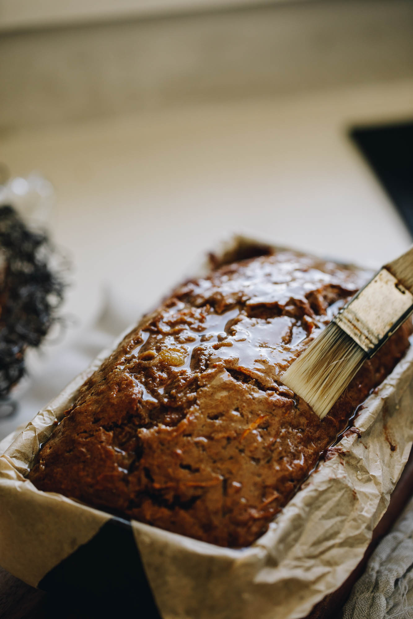 On a cream bench sits a loaf tin lined with brown baking paper. In it is a freshly baked carrot cake loaf that is being basted with pineapple juice with a pastry brush. The top is glossy. 