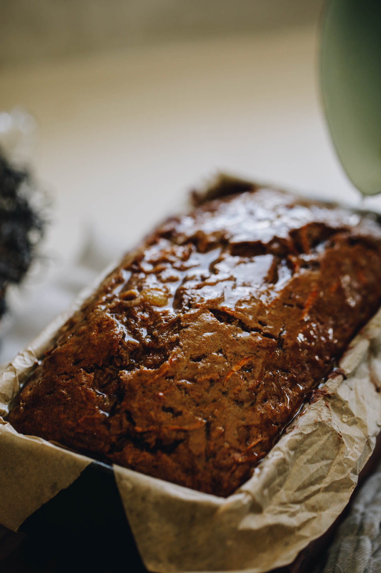 On a cream bench sits a loaf tin lined with brown baking paper. In it is a freshly baked carrot cake loaf that has been basted with pineapple juice. It is super glossy. 