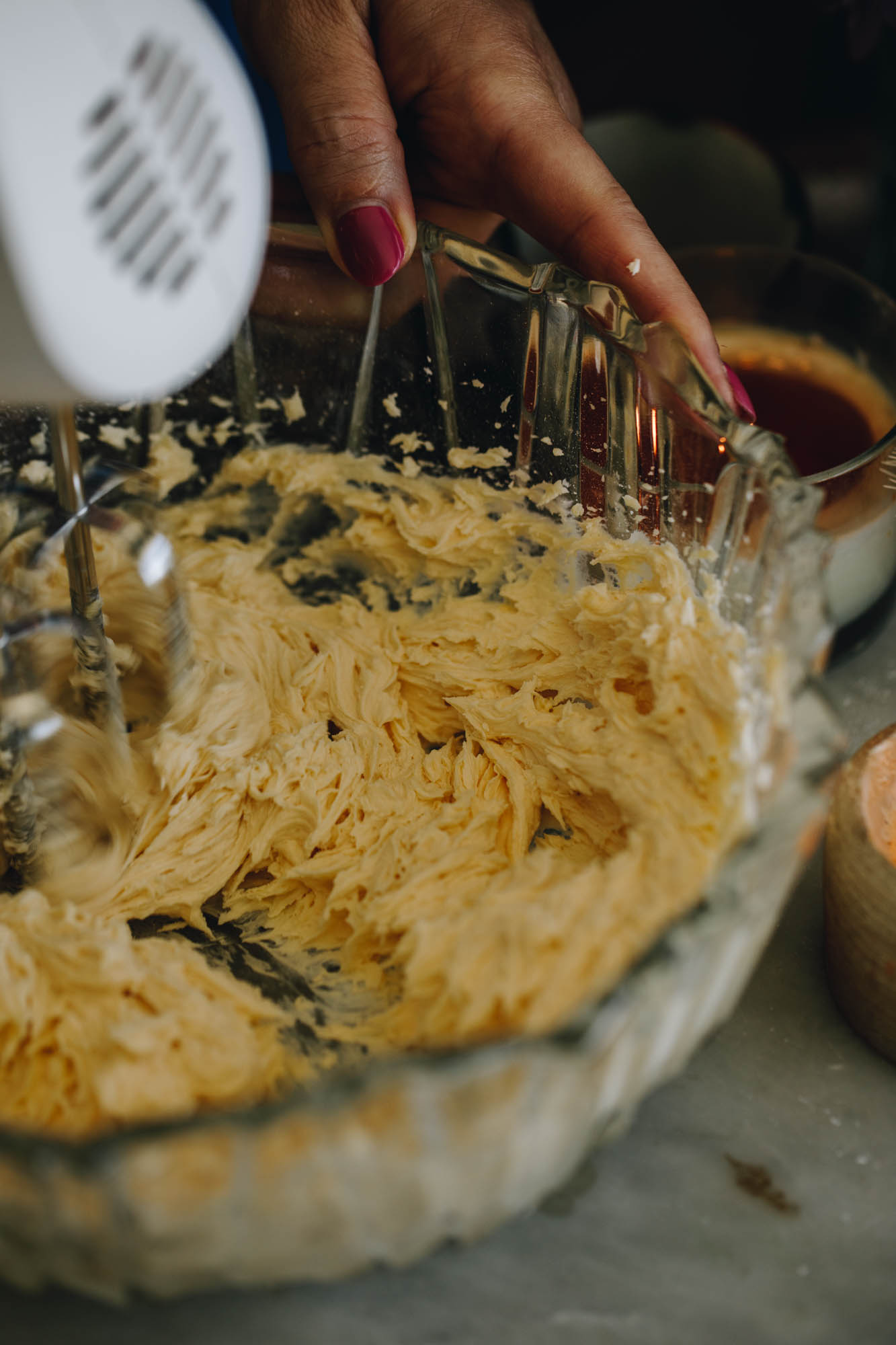 In a vintage glass bowl is butter and sugar being whipped with a w white hand mixer. It is sitting on a green tablecloth. 