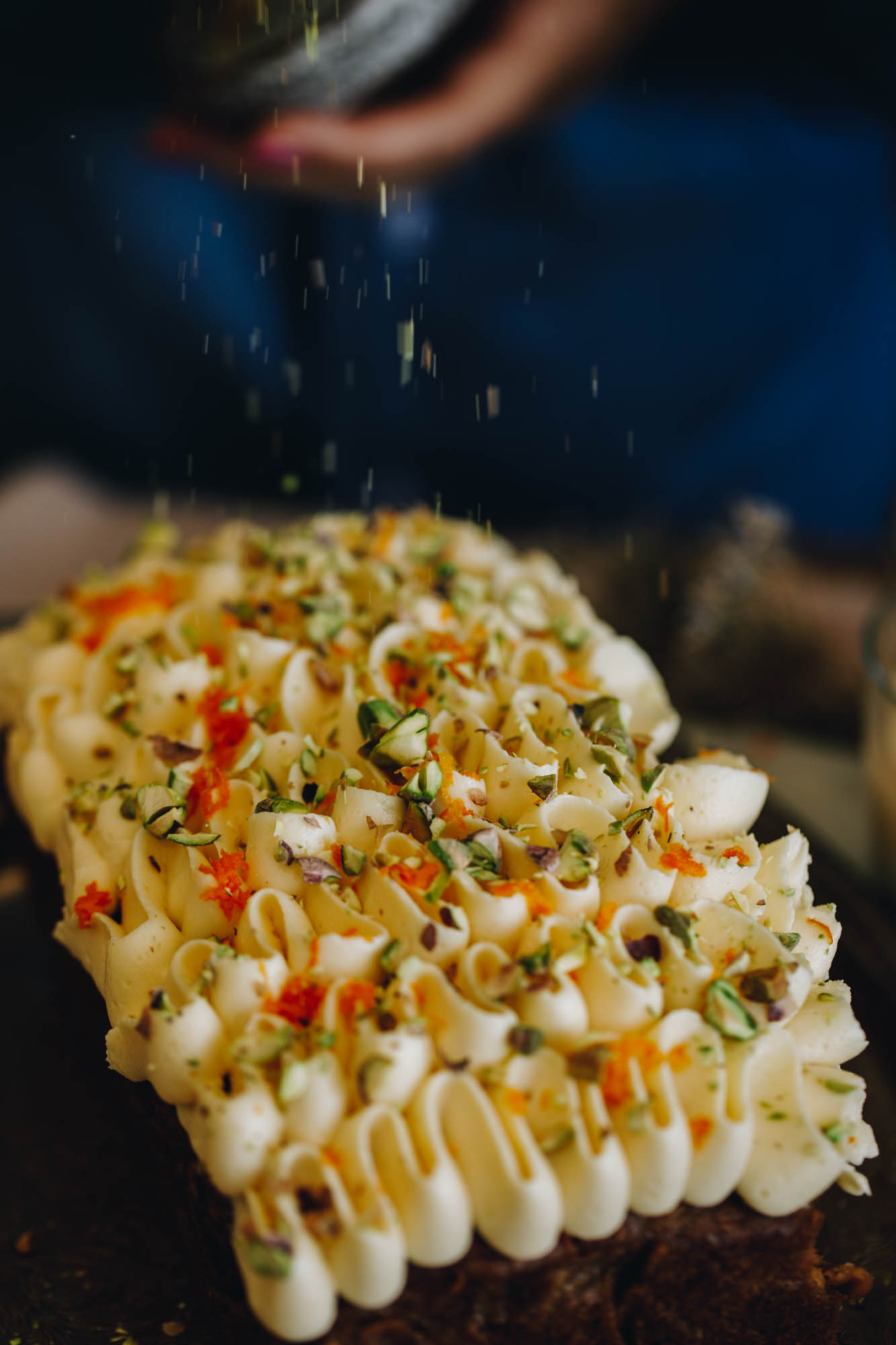 The Carrot Cake Loaf is baked and is decorated with cream cheese frosting. An action shot shows pistachio crumb being sprinkled on top of it. Orange zest is also visible. 