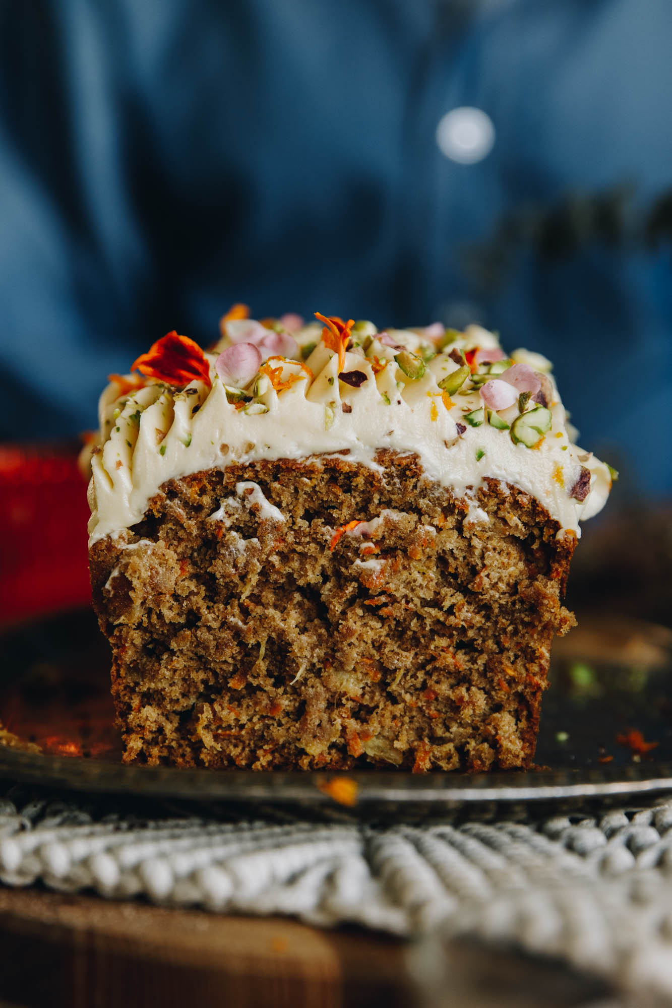 On a wooden table sits a cream weaved mat. On the mat is a freshly bake and decorated Carrot Cake Loaf that has been cut open. It has been decorated with swirls of cream cheese frosting and is topped with chopped pistachios and edible flower petals.