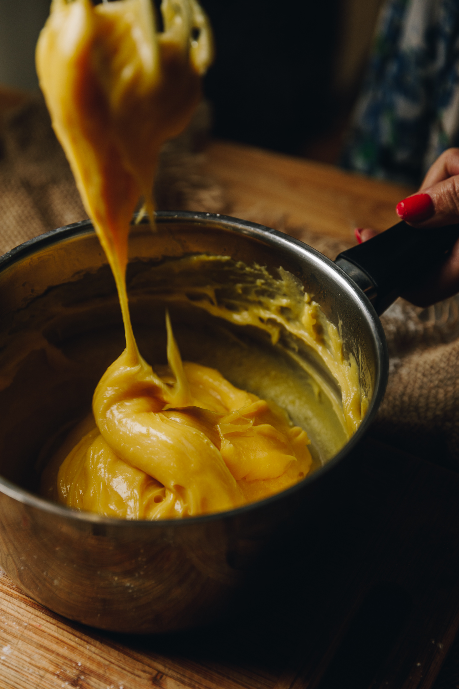 On a wooden table is a pot. Inside is thick custard that is being lifted out showing texture. 