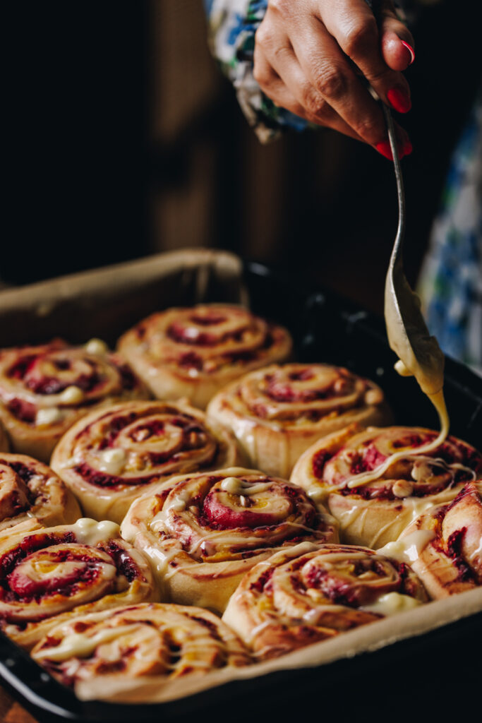A tray of baked custard and raspberry scrolls are having melted white chocolate drizzled on top of them. 