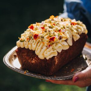 A whole Carrot Cake Loaf has been decorated with cream cheese frosting and small flower petals and chopped pistachios. The loaf is on a small tray.