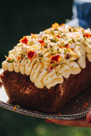 A whole Carrot Cake Loaf has been decorated with cream cheese frosting and small flower petals and chopped pistachios. The loaf is on a small tray.