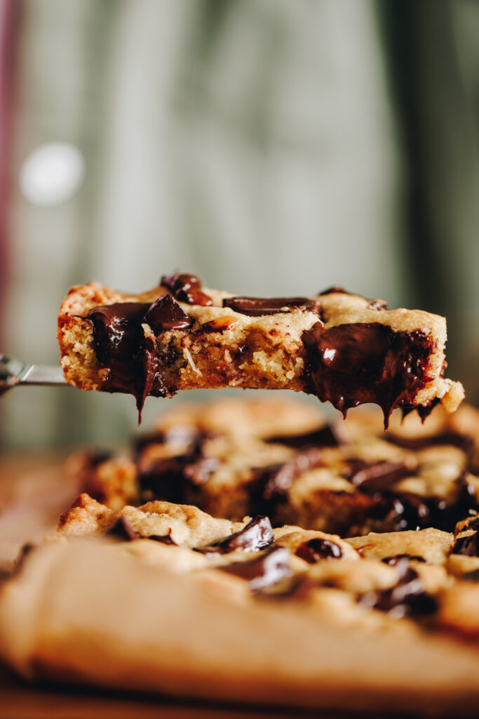 A pistachio chocolate cookie slice is held up in the air revealing the gooey chocolate centre. 