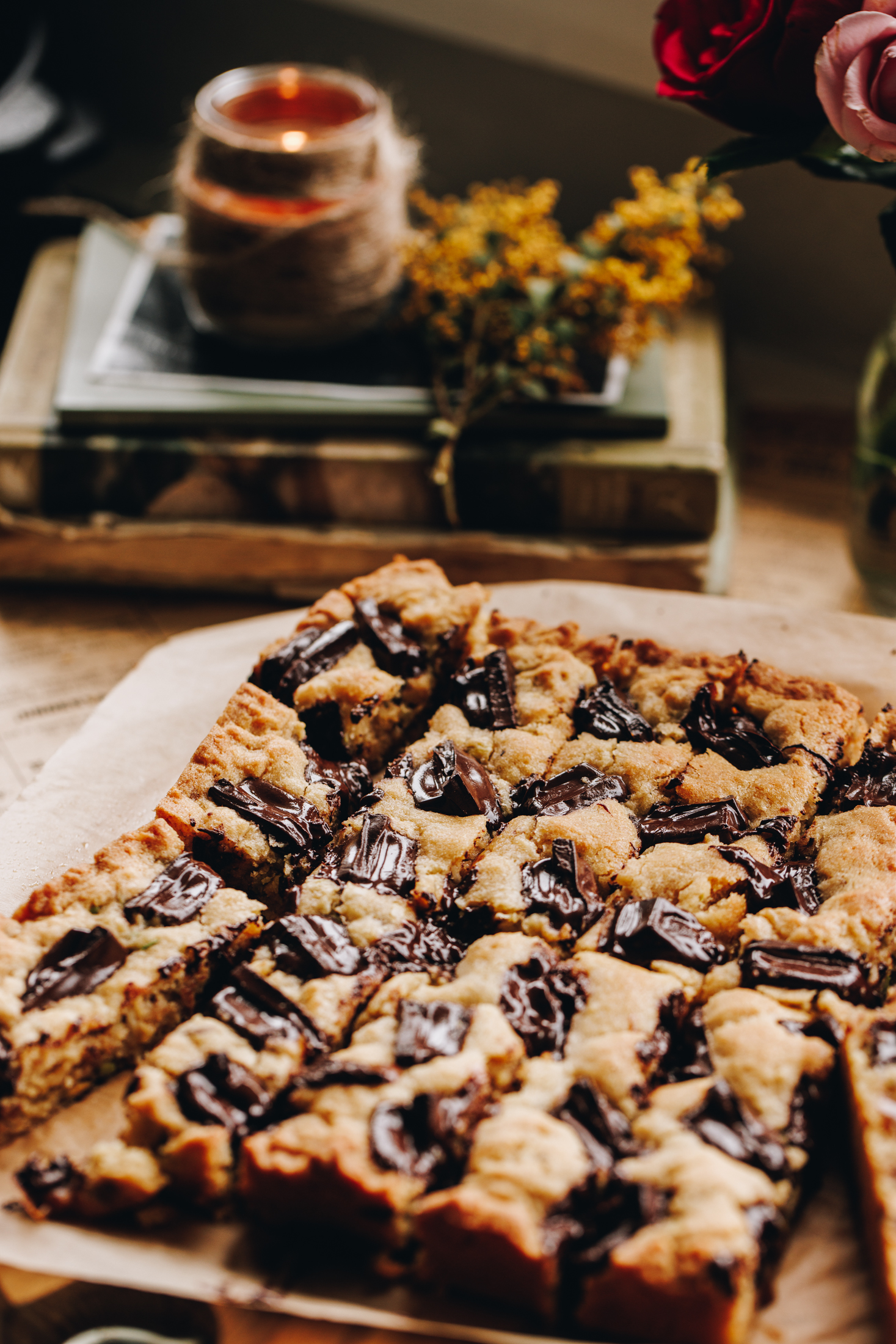 A whole pistachio chocolate cookie slice is on a wooden board and is sliced in to long pieces. Books, candles and flowers are in the background. 