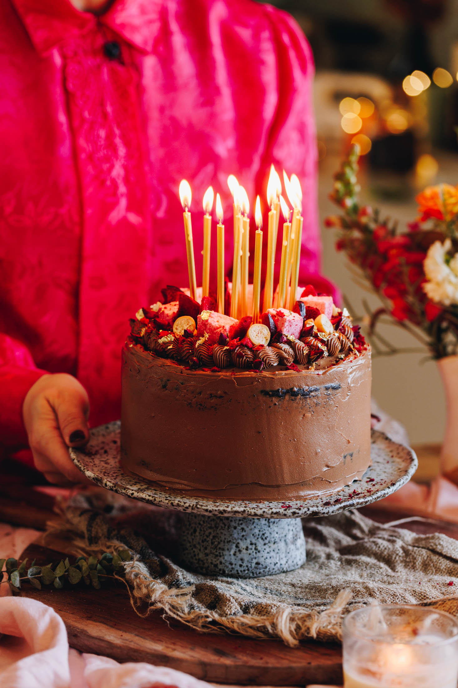On a ceramic cake stand sits a chocolate cake with biscoff buttercream. There are candles on the top that have been lit and Naomi is holding the cake stand behind it wearing a pink top. The cake is on a wooden table with flowers in the background. 
