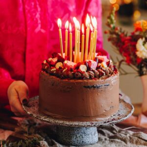 The Chocolate Cake with Biscoff Buttercream served on a ceramic cake stand with Naomi holding it from behind the table. The cake has golden candles that have been lit for celebration on it.