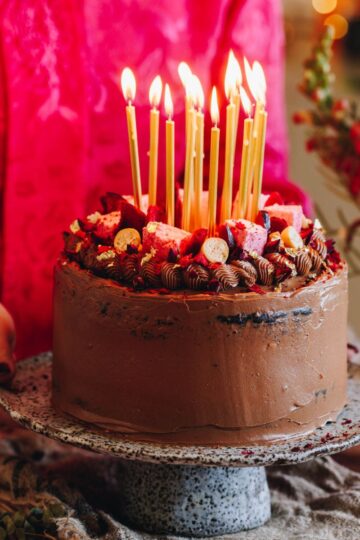 The Chocolate Cake with Biscoff Buttercream served on a ceramic cake stand with Naomi holding it from behind the table. The cake has golden candles that have been lit for celebration on it.