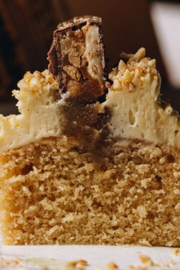 A Brown Butter Vanilla Cupcake has been sliced open to reveal the cake, gooey caramel centre, cream cheese frosting and snickers top.