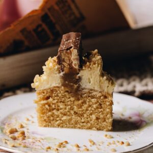 A shot of a Brown Butter Vanilla Cupcake reveals a cupcake cut down the centre revealing the soft sponge cake, gooey caramel centre, cream cheese frosting and piece of snickers bar on top. It is on a pink plate.