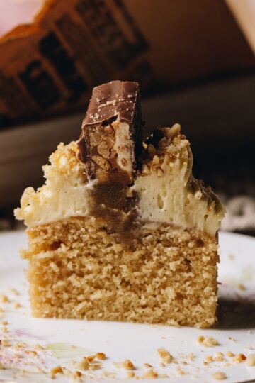 A shot of a Brown Butter Vanilla Cupcake reveals a cupcake cut down the centre revealing the soft sponge cake, gooey caramel centre, cream cheese frosting and piece of snickers bar on top. It is on a pink plate.