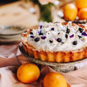 A whole Lemon Meringue Pie is on a ceramic cake stand with lemons around it, it is sitting on a pink tablecloth.