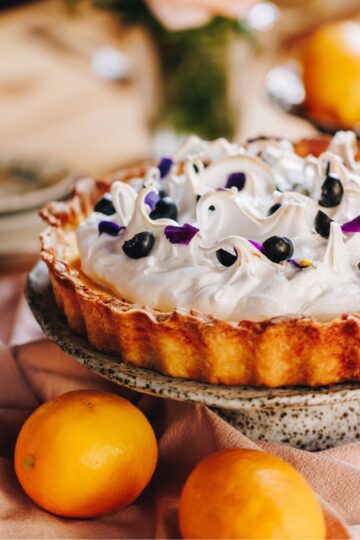A whole Lemon Meringue Pie is on a ceramic cake stand with lemons around it, it is sitting on a pink tablecloth.