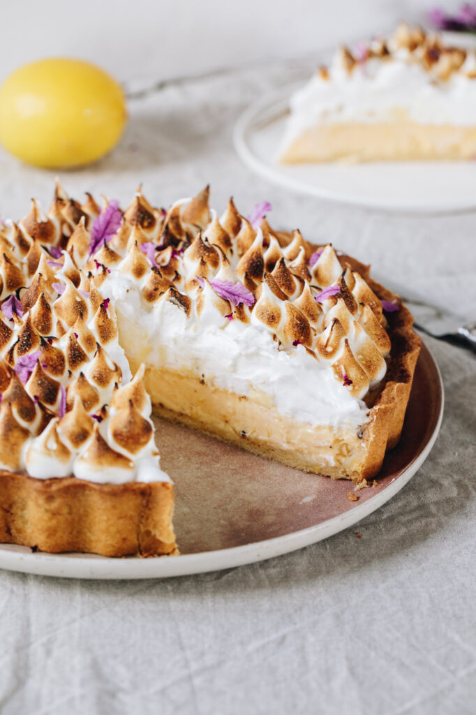 The Homemade Lemon Meringue Pie is plated on a pink plate with a lemon in the background. A large piece of pie has been removed showing the inside of the pie; the creamy lemon custard and the whispy Italian meringue that is torched. Small lavender flowers sit on top of the meringue.